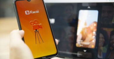 Celular com imagem de aplicativo Kwai segurado por uma mão em frente a uma tela de notebook